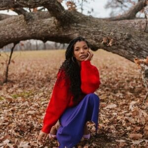 Picture of kimberly McGlonn outside during the Fall under a tree wearing a red sweatshirt and purple skirt. She is resting her head in her hand and looking directly at the camera. 