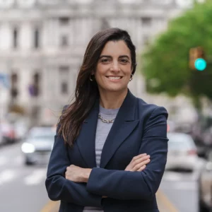 Portrait of Rebecca Rhynhart looking directly at the camera with a smile and her arms crossed. She has long brown hair and is wearing a dark blazer and light shirt. She is against a backdrop of a building and street. 