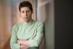 Portrait of Sam Altman with arms cross and shown from the waist up. He is wearing a lime-green long-sleeved shirt with brown hair a serious look directly at the camera. The background appears to be a nondescript office. 