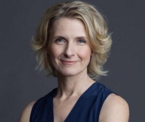 Portrait of Elizabeth Gilbert against a dark grey background wearing a blue, shoulderless v-neck and looking directly at the camera. She has medium length, wavy blond hair and is smiling. 