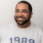 Portrait of a cheerful bearded man wearing glasses and a gray sweatshirt with the year '1989' printed in navy blue. He has short curly hair, a warm smile, and is posing against a plain, light-colored background.