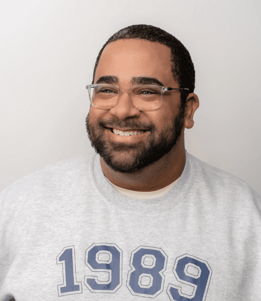 Portrait of a cheerful bearded man wearing glasses and a gray sweatshirt with the year '1989' printed in navy blue. He has short curly hair, a warm smile, and is posing against a plain, light-colored background.