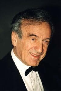 Portrait of Elie Wiesel wearing a black tuxedeo with white shirt and black bowtie shot from the waist up. Wiesel if smiling with grey-speckled brown hair.