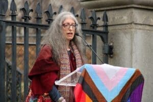 Picture of Grace Sterling Stowell speaking in front of a podium drapped in the Phildaelphia Pride flag. She has long grey hair and is wearing glasses with a red shawl and a scarf. 