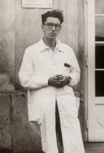 Greyscale picture of Viktor Emil Frankl with his hands together and looking directly at the camera. He has short, dark hair, wears glasses, and is wearing a white outfit. 