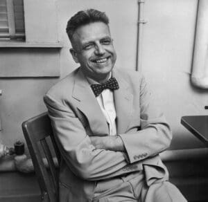 Pictured here is Alfred Kinsey in grey-scale wearinig a light blazer jacket, white shirt, and a dark bowtie. His arms are crossed and he is looking directly at the camera smiling. 