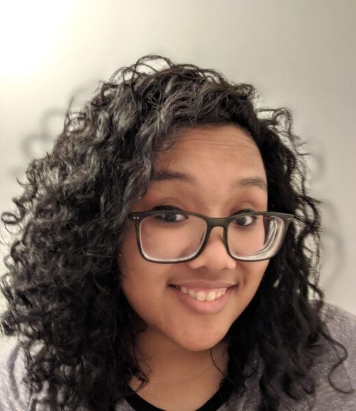 Portrait of Joanne Gonzales looking directly at the camera while smiling. She has dark, curly, and long hair and is wearing black-framed glasses. Her shirt is grey with a black collar trim.