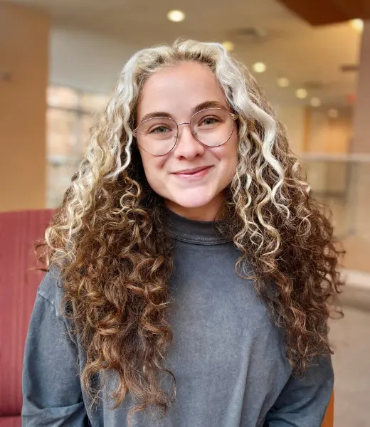 Headshot of CFR Clinical Intern Abigail Lamnin smiling at the camera indoors. She has long, curly blonde hair, round glasses, and wears a charcoal gray sweatshirt. Background is softly blurred with warm lighting.
