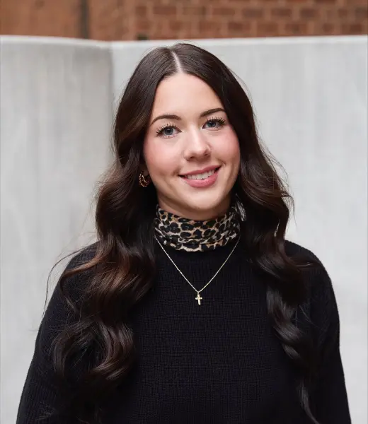 Headshot of CFR Clinical Intern Elise Rubert smiling at the camera outdoors. She has long, dark wavy hair and wears a black sweater layered over a leopard-print mock neck, gold hoop earrings, and a gold cross necklace. Background is softly blurred with a light wall and brick detail.