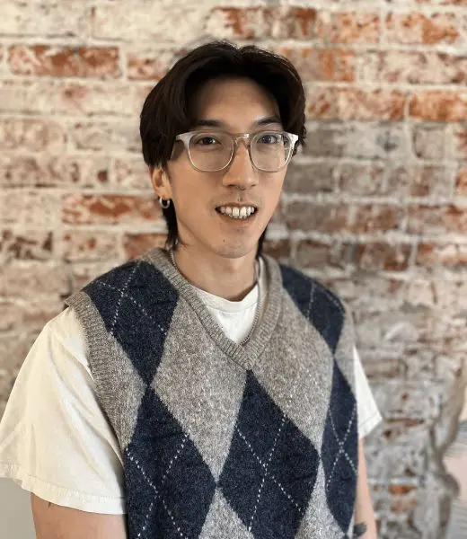 Headshot of Harim Jung smiling at the camera in front of a whitewashed brick wall. Harim has short dark hair, clear-framed glasses, and a small hoop earring, and is wearing a white T-shirt under a gray and navy argyle sweater vest.
