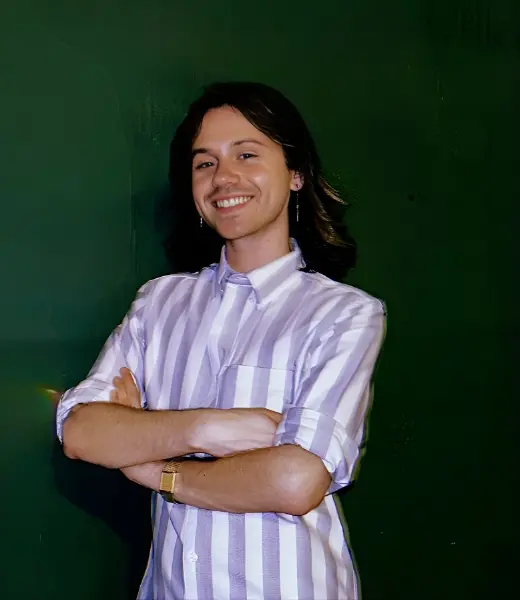 Headshot of CFR Clinical Intern Matthew Venuti smiling with arms crossed against a dark green background. They have medium-length dark hair and wear a light purple and white striped button-down shirt, earrings, and a watch.