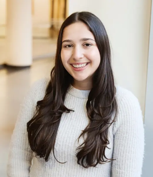 Headshot of CFR Clinical Intern Rose Stagliano smiling at the camera indoors. She has long dark hair and wears a light gray knit sweater. Background is softly blurred with warm hallway lighting.