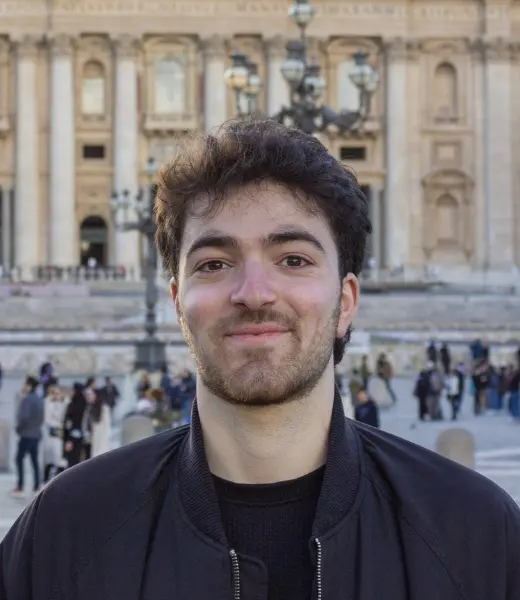 Headshot of CFR Clinical Intern Sevag Yepoyan smiling at the camera outdoors. He has dark curly hair and light facial hair and wears a dark jacket over a black top. Background shows a softly blurred historic building and people in a public square.