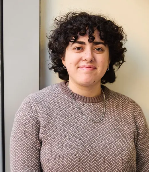 Headshot of CFR Clinical Intern Shoshana Slavitt smiling softly at the camera indoors. They have short, dark curly hair and wear a textured mauve sweater, a silver chain necklace, and a small nose ring. Background is softly blurred with warm lighting.