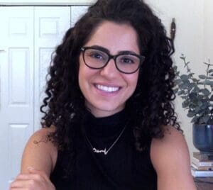 Portrait of Yasmine Beydoun looking directly at the camera and smiling. She has curly dark hair and dark-rimmed glasses. She is wearing a black, sleeveless shirt and has a gold necklace. Her arms are crossed.