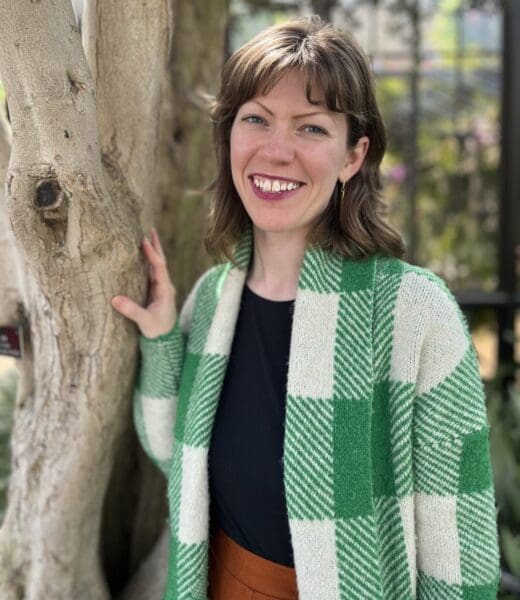Portrait of Amy Leonard looking directly at the camera against a forest backdrop. She is smiling with long, brown hair and is wearing a green and white plad button down shirt, unbuttoned, and a dark undershirt.
