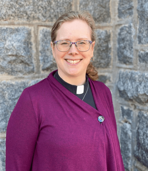 A headshot of Jennifer Phelps, a woman with a friendly smile, wearing glasses and a purple collared shirt with a clergy collar. She has her hair pulled back and is sporting a circular pin on her top. The background features an outdoor setting with a stone wall, highlighting her cheerful demeanor in a natural light.