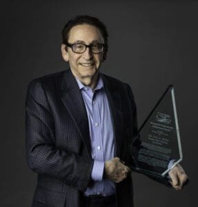 Portrait of Dr. Joel Myers holding an award smiling and looking directly at the camera. He is wearing a dark sports jacket and a blue button down shirt. He has short hair and wears dark-rimmed glasses. 