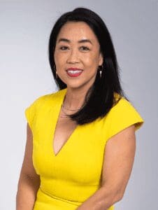 Portrait of Helen Gym against a plain grey backdrop. Gym is wearing a yellow shirt and is looking directly at the camera while smiling. She has dark hair down to her shoulders and the portrait is cut off at her waist. 