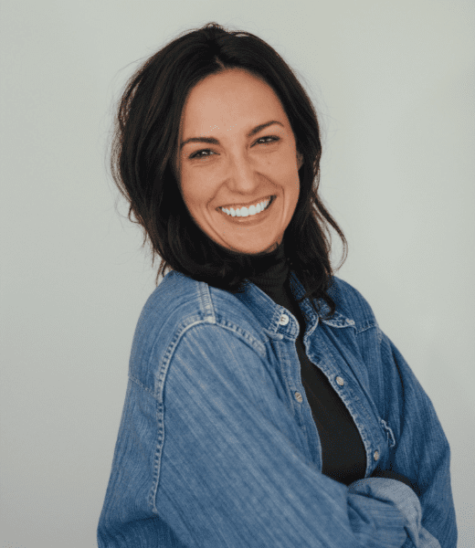 A vibrant headshot of Polly Palmer, a woman with a radiant smile, shoulder-length dark hair, wearing a dark green turtleneck and a light-wash denim jacket. She's standing with her arms crossed against a plain white background, exuding a friendly and approachable demeanor.