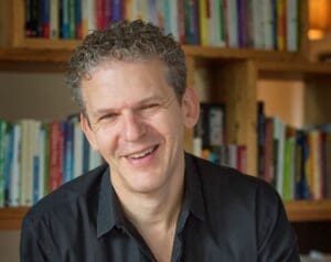 Portrait of Dr. Russ Harris in front of a bookcase. He is looking directly at the camera and is smilling with short, curly hair. He is wearing a dark shirt with the top button undone. 