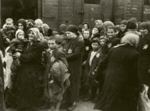A picture from The Auschwitz Album in grey scale showing a group of Jewish people of varying ages standing in front of a railroad box car. Some of the people are looking directly in the camera.