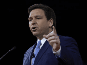 Picture of Ron DeSantis against a black background wearing a blue tie and dark blue suit. He is pointing off screen and is in the middle of speaking. 