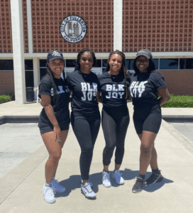 Picture of the 4 founders of Upset Homegirls standing in front of a building all looking directly at the camera and wearing black shirts that say Blk Joy.