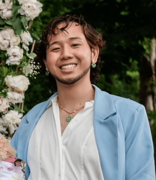 A cheerful person at an outdoor event, wearing a light blue blazer over a white shirt, complemented with green earrings and a necklace. They are smiling broadly, standing in front of a floral arrangement with white and blush flowers, which adds a festive and elegant atmosphere to the image.