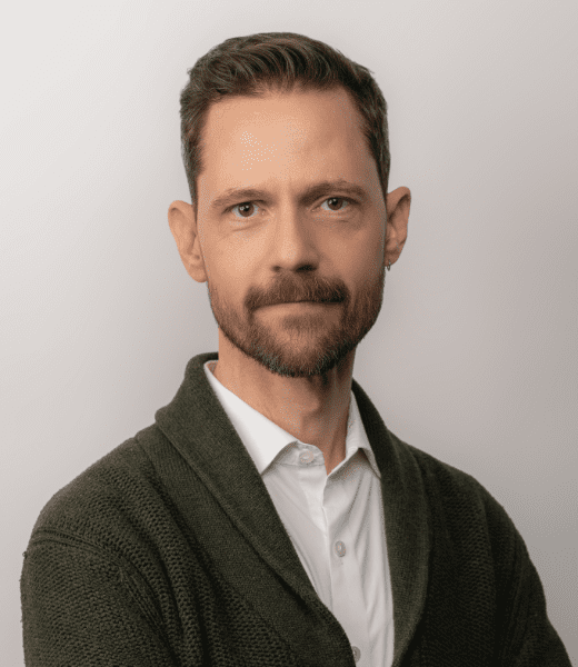 A professional headshot of Seth Fisher, a person with neatly groomed facial hair and a subtle smile, sporting a white shirt and a dark green cardigan. His direct gaze and confident posture are set against a light grey background, conveying a composed and trustworthy appearance.