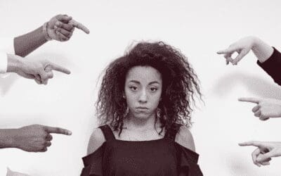 Black and white image of a young women in a dark shirt and light pants with a sad facial expression and hands in her pockets. She is surrounded by pointing fingers.