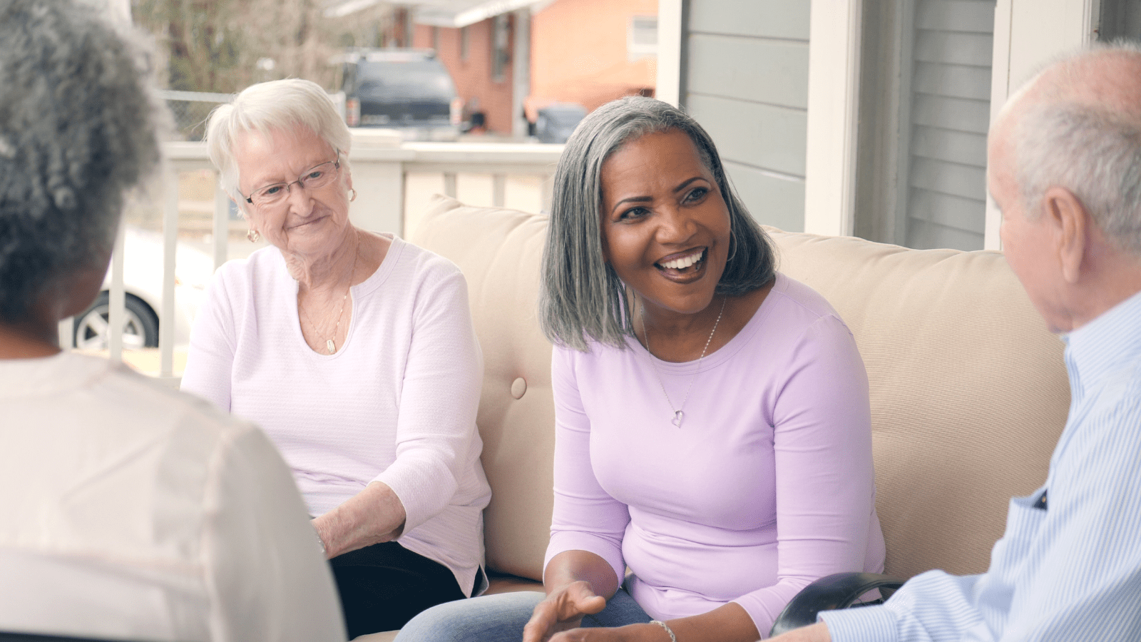 Four older adults, including two women and two men, sitting on a porch sofa, engaged in a lively conversation. One woman with gray hair in a purple top is smiling brightly, while the others listen attentively, creating a warm and friendly atmosphere