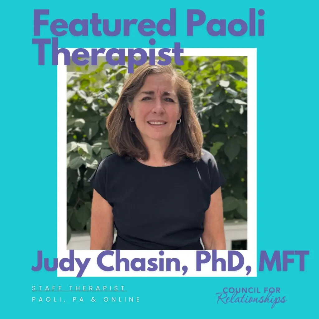 A professional photo of Judy Chasin, PhD, MFT, featured as a Paoli-based staff therapist at Council for Relationships. She is wearing a black top and standing outdoors in front of green foliage. Text on the image reads: "Featured Paoli Therapist – Judy Chasin, PhD, MFT – Staff Therapist, Paoli, PA & Online." Judy offers professional help to individuals and couples navigating burnout, grief, and symptoms of depression. She supports clients asking themselves questions like "how do you know you need therapy?" and "how to know if someone is depressed?" Judy provides therapy for grief and specializes in helping clients access expert, compassionate care.