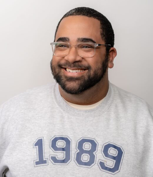 A man with a warm smile is pictured in a headshot. He has closely cropped hair and a neatly trimmed beard. He is wearing clear-framed glasses and a casual, heather gray crewneck T-shirt with the number "1989" printed in a large, varsity font. The background is plain and light-colored, putting the focus on his friendly demeanor.