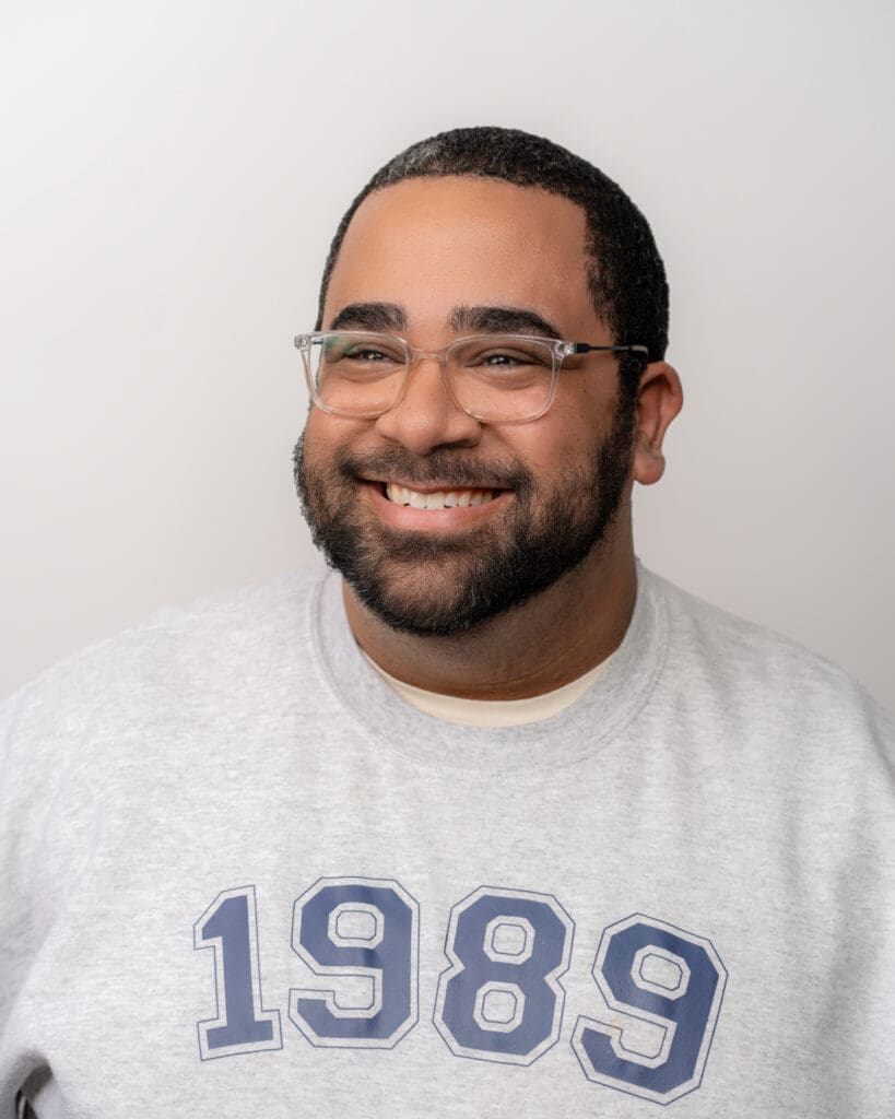 A man with a warm smile is pictured in a headshot. He has closely cropped hair and a neatly trimmed beard. He is wearing clear-framed glasses and a casual, heather gray crewneck T-shirt with the number "1989" printed in a large, varsity font. The background is plain and light-colored, putting the focus on his friendly demeanor.