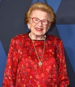 Picture of Ruth Westheimer wearing a red shirt with gold necklace and red glasses. She is looking directly at the camera and is smiling. 