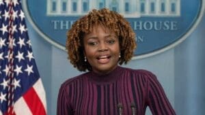 Picture of Karine Jean-Pierre standing the in White House Briefing Room wearing a purple shirt and looking directly at the camera while speaking. 