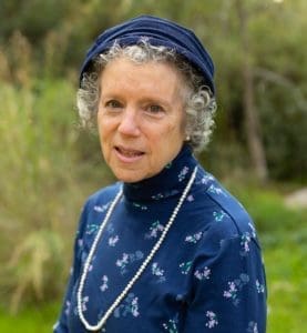 Profile of Dr. Nancy Isserman with her left shoulder forward and looking directly at the camera against a green, natural background. She is wearing a blue turtleneck with a white necklace. She has short hair is a smiling. She is also wearing a blue hat.
