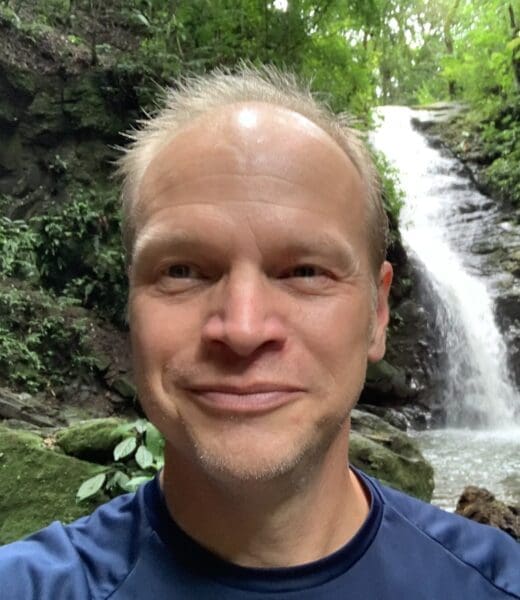A man with a content smile is pictured in a selfie, with a waterfall cascading in the background surrounded by lush greenery. He has short, light hair, and is wearing a navy blue top. The setting appears to be a serene, forested area, suggesting a moment of enjoyment in nature.