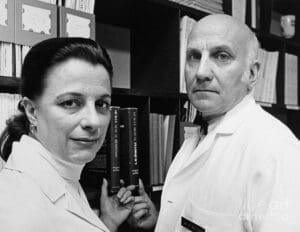 Picture of William Masters (right) and Virginia Johnson (left) in grey scale standing in front of a book case. Both Masters and Johnson are wearing white shirts and looking directly at the camera with neutral expressions.